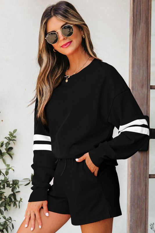 Woman in black outfit with white stripes, wearing sunglasses, standing near a window and plants.
